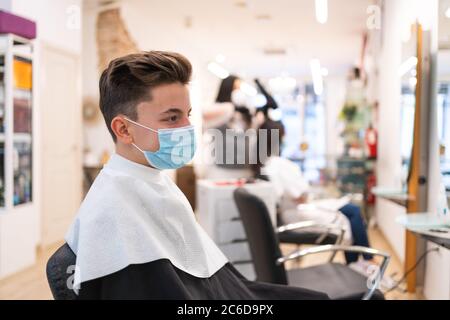 Ritratto di profilo di un ragazzo con una maschera protettiva seduta su una sedia in un parrucchiere vestaglia e un parrucchiere asciugando i capelli a un cl Foto Stock