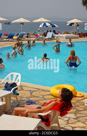 I vacanzieri, principalmente donne, si divertono in una lezione di ginnastica in piscina presso un hotel, Corfù, Grecia, e altri seduti intorno a rilassarsi al sole Foto Stock