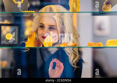 Un giovane, bella donna bionda guarda gioielli in ambra presso un negozio gioielli. Vista attraverso il vetro dal lato della strada - immagine Foto Stock