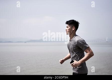 giovane adulto asiatico che corre jogging all'aperto sul mare, vista laterale Foto Stock