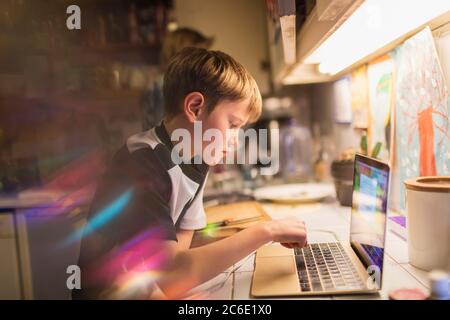 Ragazzo focalizzato che fa i compiti al computer portatile in cucina Foto Stock
