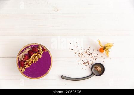 Frullato Acai, muesli, semi, bacche fresche in una ciotola di legno. Piatto riempito con bacche e cucchiaio di ceramica su fondo di legno bianco. Overhead Foto Stock