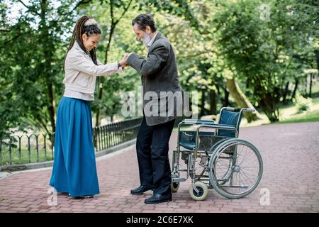 Anziano disabile che cerca di alzarsi dalla sedia a rotelle, con l'aiuto della sua bella giovane nipote con lunghi capelli temerati, sorridente e. Foto Stock