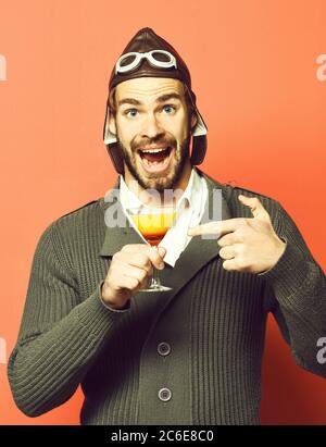 bell'uomo aviatore con bearded e barba lunga sul volto sorridente che tiene un bicchiere di cocktail alcolico in maglia grigia con cappello e bicchieri, su sfondo rosso dello studio Foto Stock
