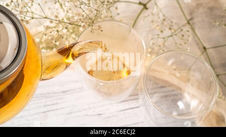 Dalla teiera in vetro trasparente versare il tè dorato in una tazza di vetro. Teiera di vetro che versa il tè nero nella tazza. Tavolo di legno bianco Foto Stock