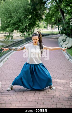 Ritratto di carismatica ragazza con dreadlock, con gonna lunga blu e t-shirt bianca, che si posa a macchina fotografica in piedi in un vicolo del parco cittadino con le braccia Foto Stock