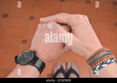 Uomo che indossa un bracciale in corda, legno, pelle, perline e sacco di corda colorato. Orologio nero sulla mano dell'uomo. Moda Hipster su pavimento piastrellato sfondo Foto Stock