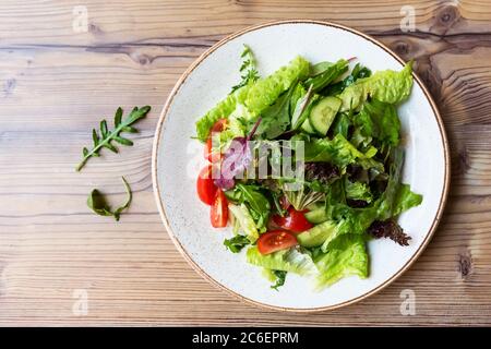 Insalata di verdure fresche con pomodoro, cetriolo, cipolla, spinaci, lattuga e rucola su piatto bianco. Ciotola di insalata con verdure su tavola di legno. Foto Stock