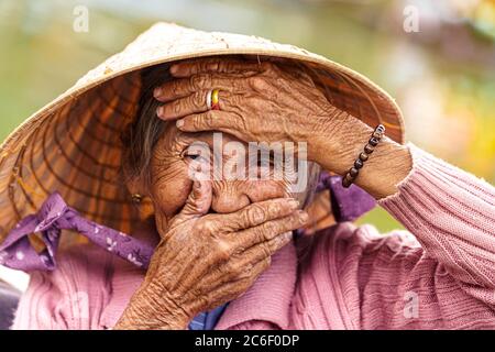 La vecchia donna vietnamita in felpa viola le mette le mani sul viso per coprire la bocca e la fronte. Foto Stock