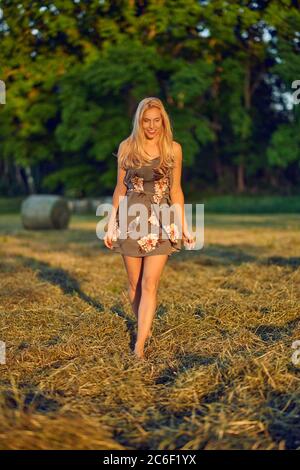 Bella donna bionda snella in un campo di fattoria che cammina verso la macchina fotografica in un abito estivo alla moda nel caldo bagliore della luce della sera Foto Stock