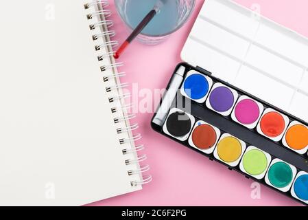 vista sopra la paintbox acquerello, pennello, libro di schizzi e vetro d'acqua su sfondo rosa, pittura con acquerelli e di essere creativo concetto Foto Stock