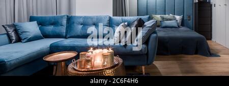 Panorama di appartamento in blu con grande divano ad angolo, tavoli da caffè in rame con decorazioni e camera da letto aperta Foto Stock