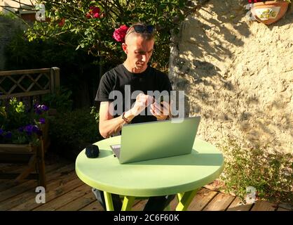 Uomo che lavora su un computer portatile a casa nel suo giardino. Lavorare per la distanza sociale. Persone che fanno shopping online o datare con Internet su smartphone. Foto Stock