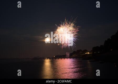 I fuochi d'artificio esplodono nel cielo sul fiume Tappahannock in Virginia. L'immagine temporizzata cattura i riflessi nell'acqua e nella Luna piena Foto Stock