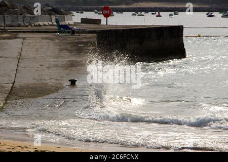 Onda che si schianta contro uno scivolo di pietra con una spiaggia di sabbia in primo piano e luce solare riflessa e due lettini vuoti Santander Cantabria Spagna Foto Stock