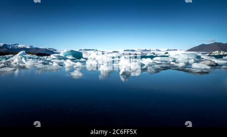 un ampio colpo di ghiaccio galleggia nelle acque fredde e calme della laguna di jökulsarlon, di fronte a un enorme ghiacciaio islandese sullo sfondo Foto Stock