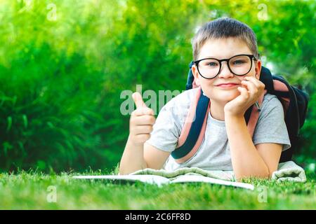 ridendo bambino bambino che mostra il pollice in su guardando alla macchina fotografica. ragazzo in occhiali con borsa di scrittura compiti nel parco. bambino maschio fa note all'aperto. Felice Foto Stock