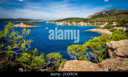 vista pittoresca attraverso i rami di pino sulla baia di sant'elm con barche al centro, l'isola di sa dragonera sul lato sinistro e le montagne Foto Stock