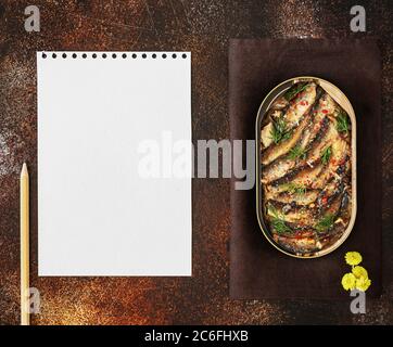 Aprire lo stagno di spratti baltici in olio con spezie con un foglio di carta da scrittura bianca e una matita su un vecchio sfondo marrone Foto Stock