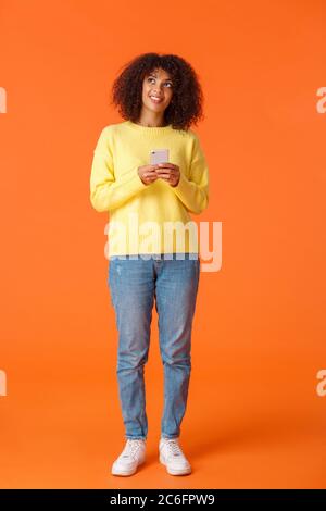 Full-length colpo verticale sognanti carino afro-donna americana pensando a cosa scrivere, tenendo lo smartphone pensare guardando verso l'alto e sorridente cose di imaging Foto Stock