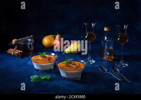 Panna cotta dessert tradizionale italiano con pere in camicia al rum e sciroppo di miele. Tavola di sfondo blu scuro, bicchieri di rum, vasetto di miele, pere e. Foto Stock