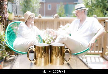 Una coppia matura seduta sul loro patio durante una bella giornata estiva. Davanti a loro su un piccolo tavolo, hanno due tazze di caffè dorate. Foto Stock