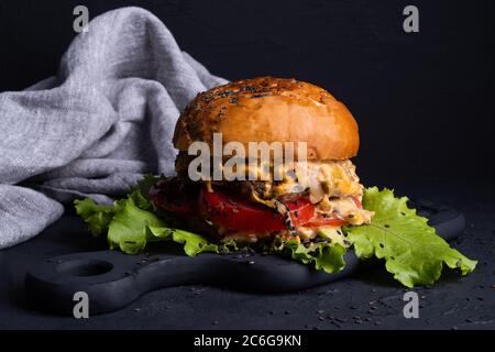 hamburger grande con verdure, manzo e salsa su fondo nero, sullo sfondo di tovagliolo grigio, salsa proveniente da hamburger, pomodori e fresco Foto Stock