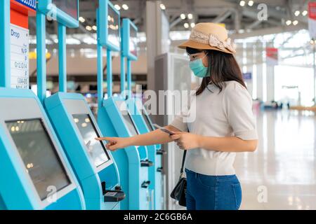 Donna asiatica che indossa una maschera utilizzando un chiosco di check-in self-service nel terminal dell'aeroporto durante la prevenzione delle pandemie del coronavirus (COVID-19) quando si viaggia all'estero. Foto Stock