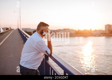 Un uomo d'affari caucasico di mezza età con occhiali, in una camicia bianca, che parla sul telefono appoggiandosi contro la ringhiera del ponte Foto Stock