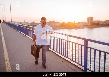 Un uomo d'affari caucasico di mezza età con occhiali, in una camicia bianca e con una valigetta nera in mano cammina sul ponte e guarda il suo orologio Foto Stock