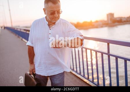 Un uomo d'affari caucasico di mezza età con occhiali, in una camicia bianca e con una valigetta nera in mano cammina sul ponte e guarda il suo orologio Foto Stock