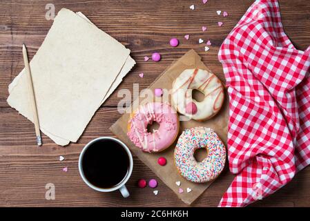 Ciambelle con spolverare i cuori, tazza di caffè e vecchio foglio di carta, sfondo di legno, vista dall'alto Foto Stock