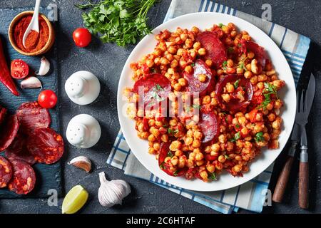 Primo piano di Garbanzos fritos, stufato di ceci con chorizo a fette, prosciutto, pomodori e spezie su un piatto bianco su un tavolo di cemento con ingredienti, s. Foto Stock