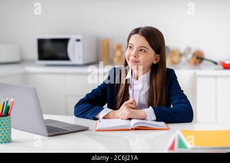 Foto di piccola bella scuola studentesca ragazza notebook tavolo lezione online videochiamata scrivere note saggio guardare sognante in su pensare la distanza Foto Stock