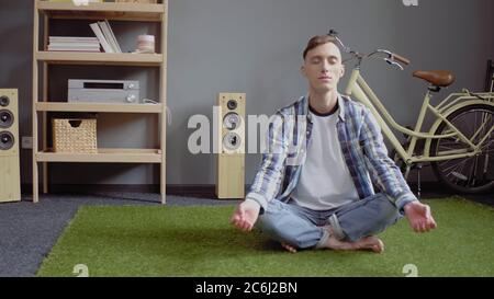 L'uomo caucasico medita gli occhi chiusi mentre si siede le gambe incrociate su un tappeto verde. Autoisolamento a casa. Concetto di quarantena. Immagine a toni Foto Stock