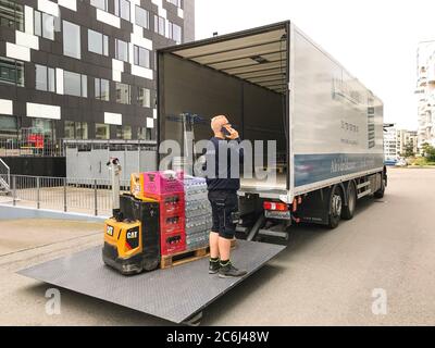 Il delivery man parla su un telefono cellulare mentre le bevande vengono scaricate con un carrello elevatore a forche su un caricatore a portellone di un carrello di consegna davanti a un edificio per uffici Foto Stock