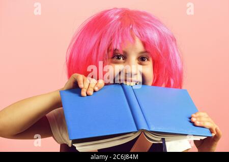 Ragazza morde grande libro blu. Ragazza della scuola con faccia felice isolata su sfondo rosa. Torna a scuola e concetto di istruzione. Alunno in uniforme scolastica con parrucca rosa. Foto Stock