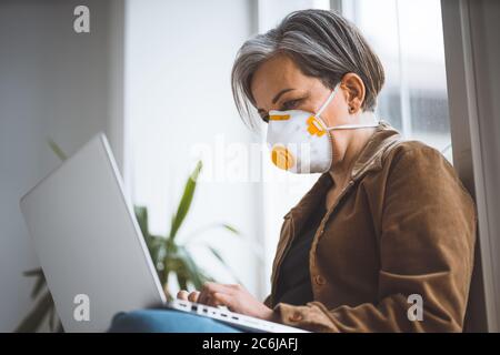 Donna con capelli grigi e maschera protettiva nei vestiti casual lavora su computer portatile seduto sul posto di lavoro con davanzale. Concetto pandemico. Vista laterale Foto Stock