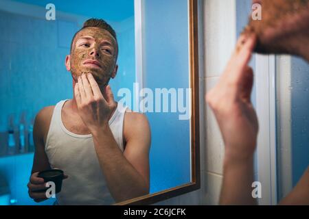 Giovane uomo che applica la maschera facciale di fronte allo specchio nel bagno domestico. Temi cura della pelle, trattamento e bellezza per gli uomini. Foto Stock