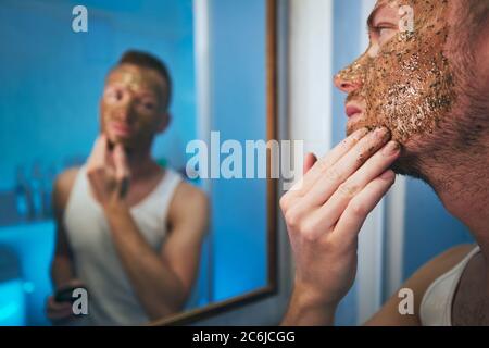 Giovane uomo che applica la maschera facciale di fronte allo specchio nel bagno domestico. Temi cura della pelle, trattamento e bellezza per gli uomini. Foto Stock