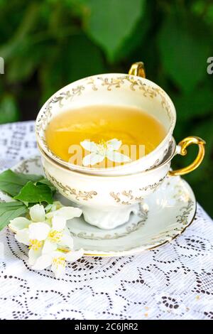 Tè al gelsomino in una tazza di porcellana con fiore di erbe di gelsomino su tela da tavolo in pizzo, sfondo foglie verdi Foto Stock