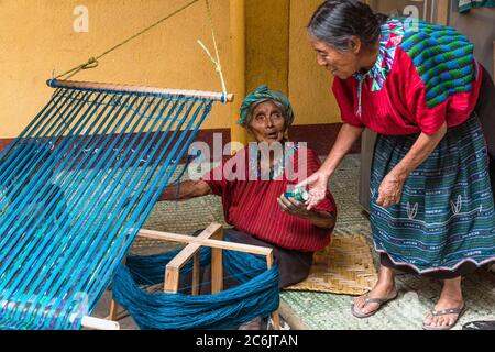 Guatemala, Solola Department, Santa Cruz la Laguna, una donna maya Cakchiquel più anziana in abito tradizionale vento filo su una cornice per prepararsi a tessitura su un telaio posteriore come il suo amico viene oltre ad aiutarla. Foto Stock