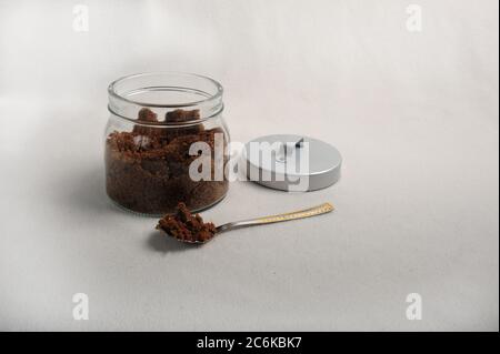 Barattolo con zucchero di canna. Un cucchiaio si trova accanto al vaso, il coperchio è appoggiato contro il vaso di vetro. Sfondo bianco, angolo di visualizzazione superiore. Spazio di copia sul lato destro Foto Stock
