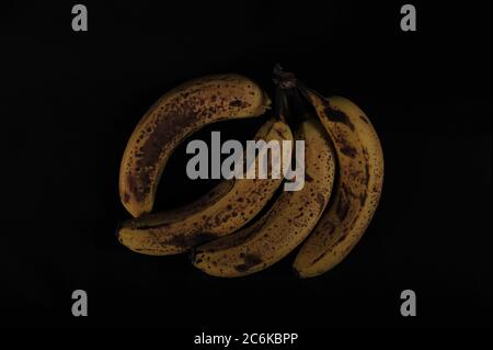 Quattro banane supermature su superficie nera. Scatto orizzontale ad angolo elevato con spazio di copia intorno ai frutti. Scatto chiave basso. Foto Stock