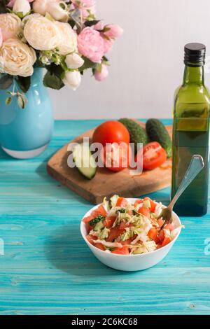 Preparazione di un'insalata vegetariana con verdure fresche e olio d'oliva. Pomodori e cetrioli sono sul tagliere, un bouquet di fiori nel backgro Foto Stock