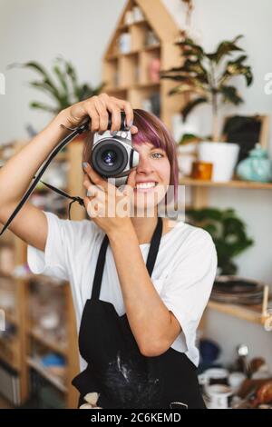 Sorridente ragazza con capelli colorati in grembiule nero scattare foto sulla macchina fotografica felicemente passare il tempo allo studio della ceramica. Foto Stock