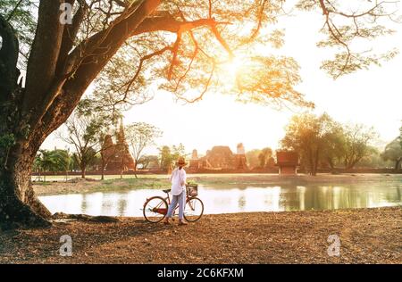 Il viaggiatore femminile ha indossato abiti estivi leggeri e cappello hanno la mattina presto a piedi con bicicletta vicino al laghetto nel parco storico Ayutthaya, Thailandia Foto Stock