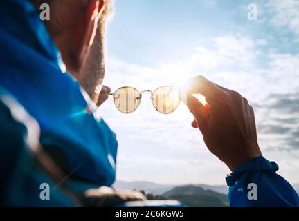 Backpacker uomo guardando il sole luminoso attraverso occhiali da sole polarizzati godendo paesaggio di montagna. Immagine del concetto di salute umana per la cura dell'occhio e della visione. Foto Stock