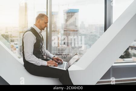 Un bel uomo d'affari maturo sta usando il laptop mentre si siede su una moderna panchina bianca vicino alla finestra di un alto ufficio di lusso Foto Stock