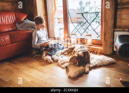 Libro di lettura del ragazzo sul pavimento vicino a scivolare il suo cane di aquila su pelle di pecora in atmosfera accogliente casa. Momenti tranquilli di accogliente casa concetto immagine. Foto Stock